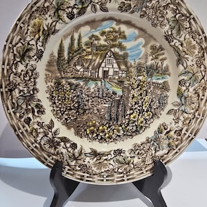 May include: A ceramic plate with a detailed landscape scene featuring a cottage, trees, and a stream. The plate has a floral border in shades of brown, green, and yellow. The plate is displayed on a black stand.
