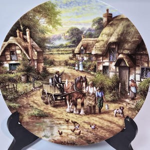 May include: A decorative plate featuring a rural village scene with thatched-roof cottages, a horse-drawn cart, and figures in period clothing. The plate is displayed on a black easel, showcasing a detailed landscape painting.