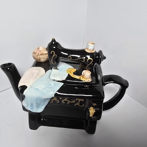 Könnte beinhalten: Schwarze Keramik-Teekanne in Form einer Nähmaschine, mit schwarzem Griff und Ausguss. Die Teekanne hat eine schwarze Nähmaschine auf der Oberseite, mit weißen und blauen Stoffakzenten und einer beigen Garnrolle.