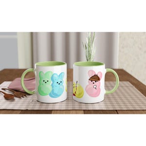 May include: Two white ceramic mugs with green handles and rims. One mug features green and blue bunny designs, the other has a pink bunny detective design. The mugs are on a checkered tablecloth.