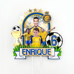 Puede incluir: Un adorno para pastel de f&uacute;tbol amarillo y azul con una foto de un ni&ntilde;o y un jugador de f&uacute;tbol. El adorno incluye un bal&oacute;n de f&uacute;tbol, un trofeo y el nombre "Enrique".