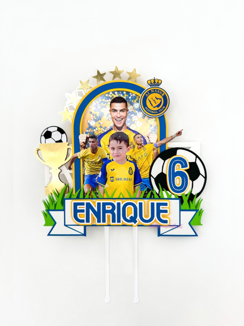 Puede incluir: Un adorno para pastel de f&uacute;tbol amarillo y azul con una foto de un ni&ntilde;o y un jugador de f&uacute;tbol. El adorno incluye un bal&oacute;n de f&uacute;tbol, un trofeo y el nombre "Enrique".