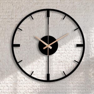 Könnte beinhalten: Eine große, kreisförmige Wanduhr mit einem schwarzen Metallrahmen, dünnen Stundenmarkierungen und einer schwarzen Mittelscheibe. Die Uhr hat zwei schlanke, goldfarbene Metallzeiger vor einer weißen Ziegelwand.