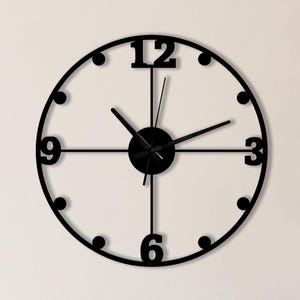 Op de afbeelding: Een zwarte, ronde wandklok met een minimalistisch ontwerp. De klok heeft zwarte wijzers en uurmarkeringen, met de cijfers 12, 3, 6 en 9 duidelijk aangegeven. De tekst "Black Hands" staat in de rechterbovenhoek. De klok is aan een lichtgekleurde muur bevestigd.