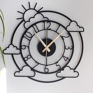 May include: A black metal wall clock with a weather theme. The clock features a sun, clouds, and a circular design with numbers. The clock hands are gold-colored. The clock is mounted on a white wall.