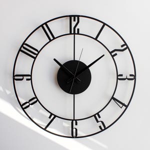 May include: A black, round wall clock with Roman numerals. The clock has a minimalist design with a black face and hands. The clock is mounted on a white wall, and the hands show the time.