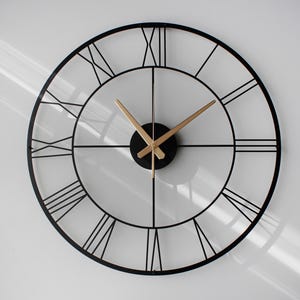 Puede incluir: Un reloj de pared grande y redondo con un marco de metal negro y números romanos. Las manecillas del reloj son doradas y el centro es un círculo negro. El reloj está montado en una pared blanca.