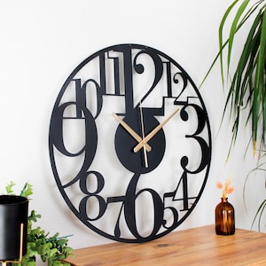 Könnte beinhalten: Schwarze Wanduhr aus Metall mit großen, fetten, überlappenden Zahlen. Die Uhr hat goldfarbene Zeiger und ein schwarzes Zifferblatt.