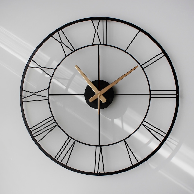 May include: A large, round wall clock with a black metal frame and Roman numeral hour markers. The clock hands are gold, and the center is a black circle. The clock is mounted on a white wall.