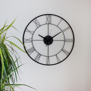 May include: A large, round, black metal wall clock with Roman numerals. The clock has a minimalist design with a black frame and hands. The clock is mounted on a white wall, with a green plant in the corner.