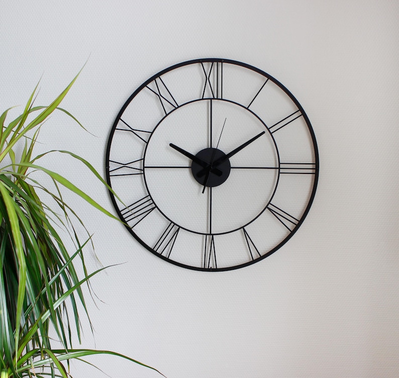 May include: A large, round, black metal wall clock with Roman numerals. The clock has a minimalist design with a black frame and hands. The clock is mounted on a white wall, with a green plant in the corner.