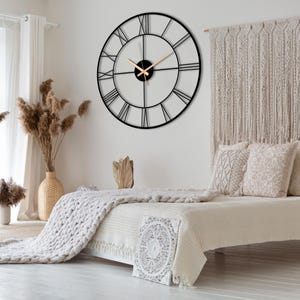 May include: A large black metal clock with Roman numeral markings on a white wall above a bed with a white knitted blanket and white pillows.