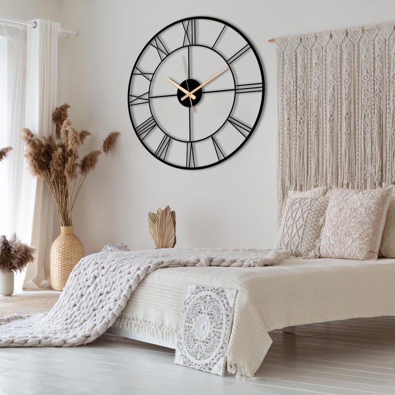 May include: A large black metal clock with Roman numeral markings on a white wall above a bed with a white knitted blanket and white pillows.