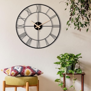May include: A large, round black metal wall clock with Roman numerals. The clock hands are light brown. The clock is mounted on a white wall. The clock's diameter is approximately 24 inches.