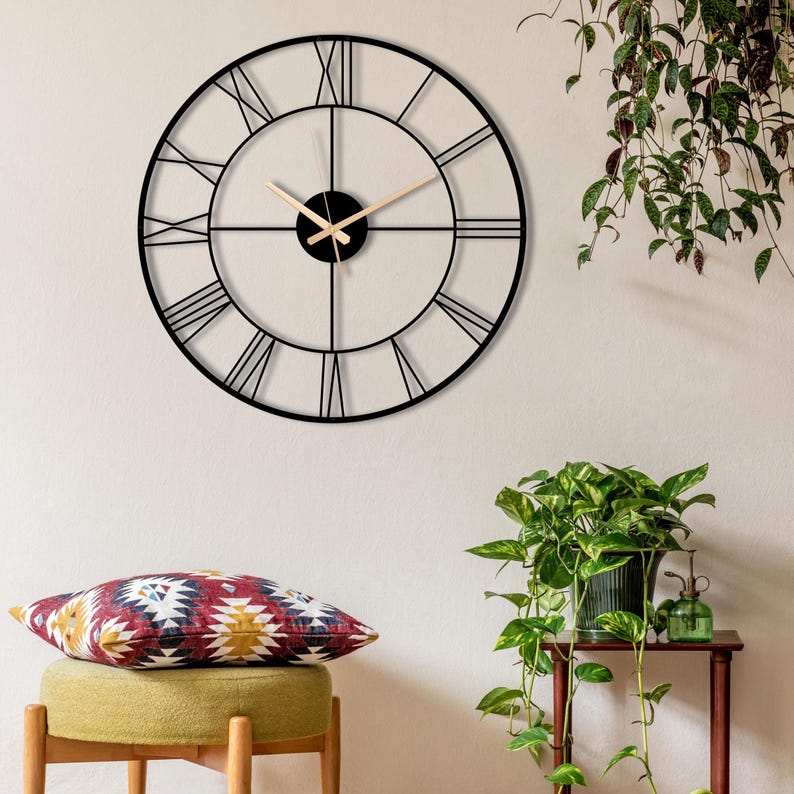 May include: A large, round black metal wall clock with Roman numerals. The clock hands are light brown. The clock is mounted on a white wall. The clock's diameter is approximately 24 inches.