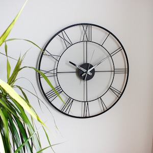 May include: A black metal wall clock with Roman numerals. The clock has a minimalist design with a circular frame and a white background. The hands are black and silver. The clock is mounted on a white wall.