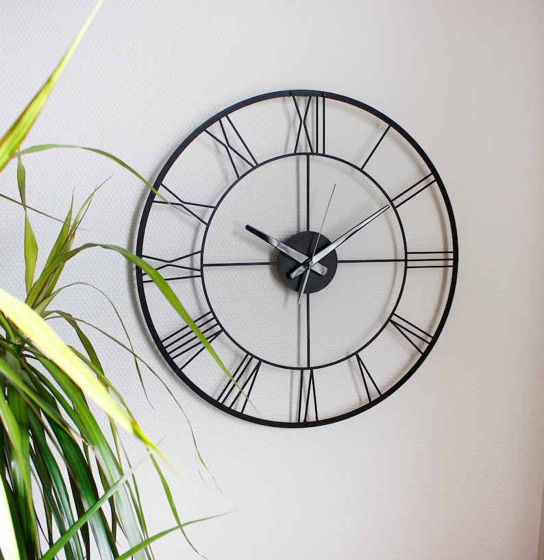 May include: A black metal wall clock with Roman numerals. The clock has a minimalist design with a circular frame and a white background. The hands are black and silver. The clock is mounted on a white wall.