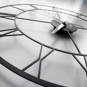 May include: A close-up of a modern, black metal wall clock. The clock face is a light gray color, with black Roman numerals and hands. The clock's design is minimalist and geometric, with a focus on lines and shapes.