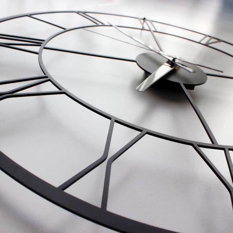 May include: A close-up of a modern, black metal wall clock. The clock face is a light gray color, with black Roman numerals and hands. The clock's design is minimalist and geometric, with a focus on lines and shapes.