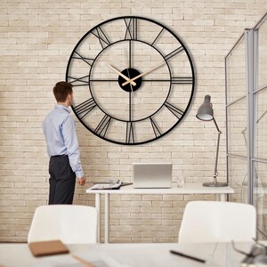 May include: A large black metal clock with Roman numeral markings on a white brick wall. The clock has a gold-colored hour and minute hand.