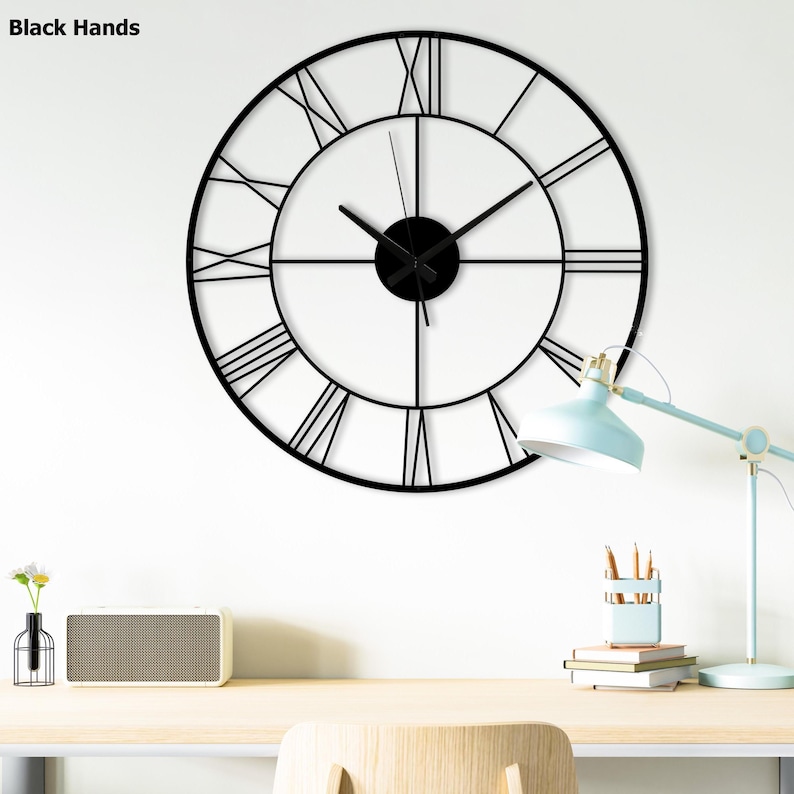 May include: A large, black metal wall clock with Roman numerals. The clock has a circular design with a minimalist aesthetic. The hands and center of the clock are also black. The clock is mounted on a white wall.