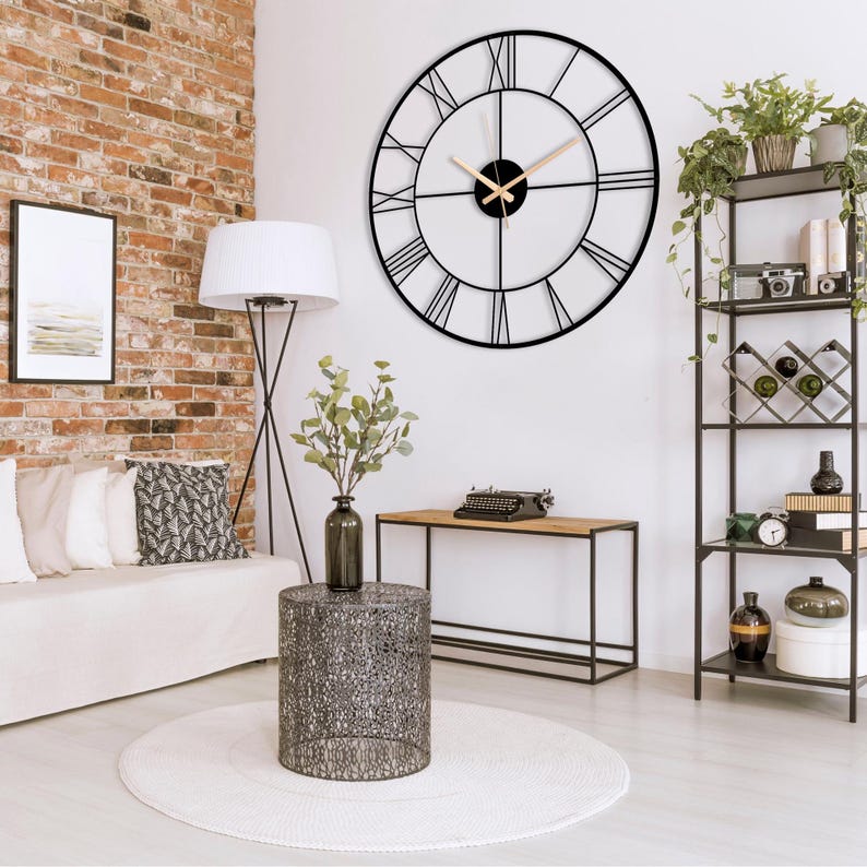 May include: A black metal wall clock with Roman numeral markings and gold-colored hands. The clock is hanging on a white wall in a living room setting with a brick wall, a white couch, a black side table, and a black console table.