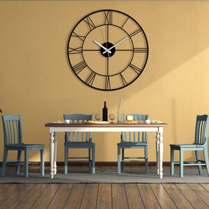 May include: A large, black metal wall clock with Roman numerals hangs above a dining table. The clock's hands are white. The dining room features a wooden table, blue chairs, and a yellow wall.
