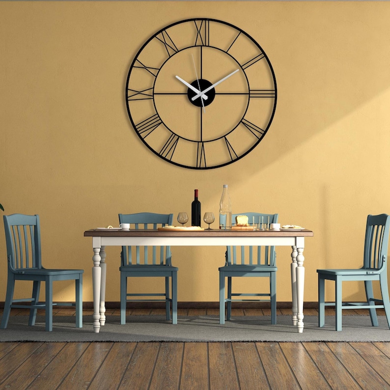 May include: A large, black metal wall clock with Roman numerals hangs above a dining table. The clock's hands are white. The dining room features a wooden table, blue chairs, and a yellow wall.