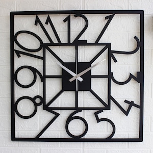 Peut inclure: Une horloge murale carrée dotée d'un cadre en métal noir avec de grands chiffres découpés de un à douze intégrés au design. Le centre se compose d'un carré plus petit avec des lignes géométriques et deux fines aiguilles de couleur argentée.