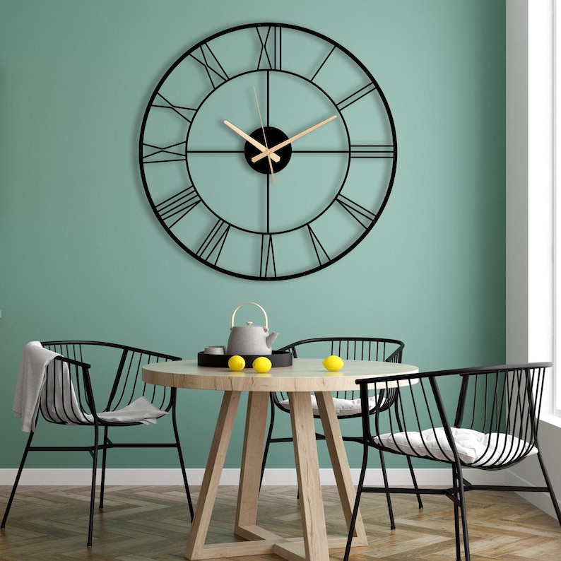 May include: A black metal clock with Roman numerals on a light green wall. The clock is hanging above a wooden table with four black metal chairs. There are lemons on the table.