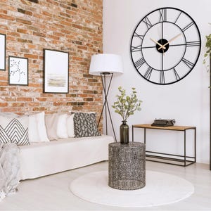 May include: A large, black metal wall clock with Roman numerals hangs on a white wall. The clock's hands are light brown. The room features a sofa with pillows, a side table, and a brick wall, creating a modern interior design.