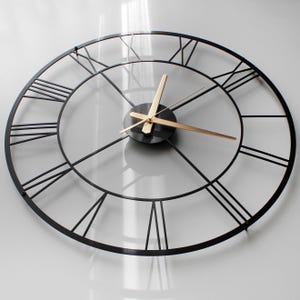 May include: A large, round, black metal wall clock with Roman numerals. The clock has a minimalist design with gold-colored hands and a black center. The clock is mounted on a white wall.
