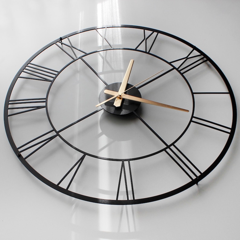 May include: A large, round, black metal wall clock with Roman numerals. The clock has a minimalist design with gold-colored hands and a black center. The clock is mounted on a white wall.