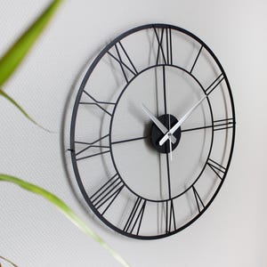 May include: A large, round, black metal wall clock with Roman numerals. The clock has a minimalist design with a black frame and silver hands. The clock is mounted on a white wall, with a plant in the foreground.