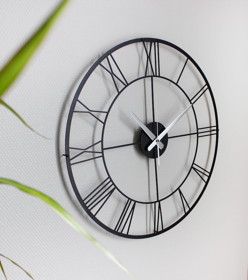 May include: A large, round, black metal wall clock with Roman numerals. The clock has a minimalist design with a black frame and silver hands. The clock is mounted on a white wall, with a plant in the foreground.