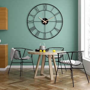 May include: A large, black metal wall clock with Roman numerals hangs above a round wooden table. The clock has a minimalist design with a black frame and light-colored hands. Three black metal chairs surround the table, creating a modern dining area.