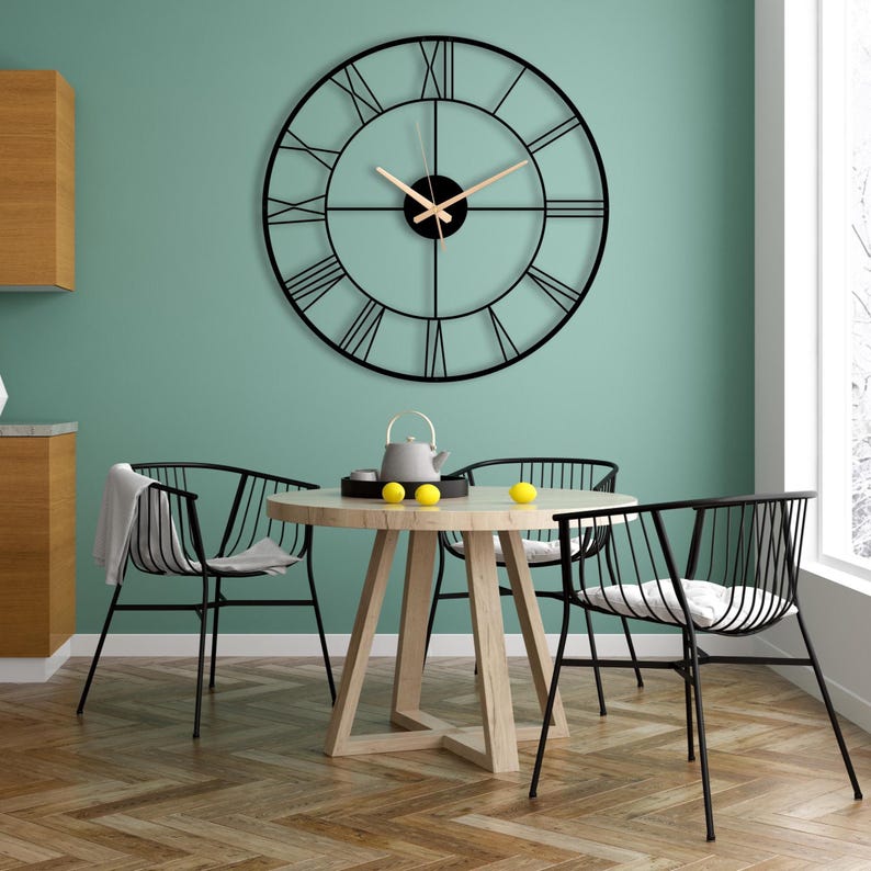 May include: A large, black metal wall clock with Roman numerals hangs above a round wooden table. The clock has a minimalist design with a black frame and light-colored hands. Three black metal chairs surround the table, creating a modern dining area.