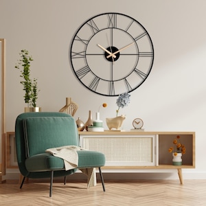 May include: A large, round black metal wall clock with Roman numerals and light-colored hands. The clock is mounted on a neutral-colored wall above a wooden cabinet and a green armchair. Decorative items like vases and a small clock are on the cabinet.