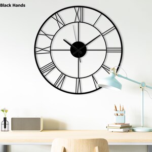 May include: Black metal wall clock with Roman numeral markings and black hands. The clock is hanging on a white wall above a wooden desk with a lamp, books, and a speaker.