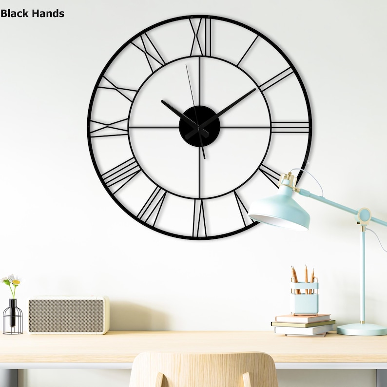 May include: Black metal wall clock with Roman numeral markings and black hands. The clock is hanging on a white wall above a wooden desk with a lamp, books, and a speaker.