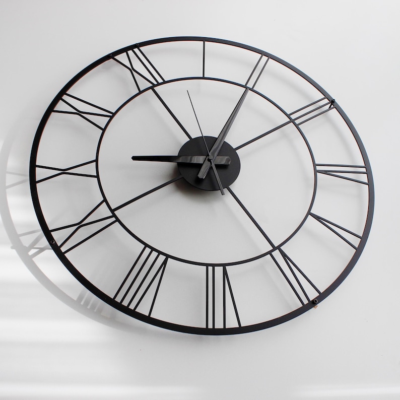 May include: A large, round, black metal wall clock with Roman numerals. The clock has a minimalist design with a circular frame and black hands. The clock's diameter is approximately 30 inches.