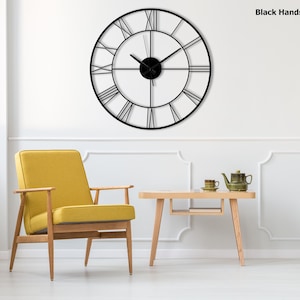 May include: A large, round black metal wall clock with Roman numerals. The clock has black hands and is mounted on a white wall. The text "Black Hands" is visible in the upper right corner. The clock is above a yellow chair and a small wooden table.