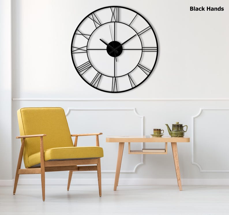 May include: A large, round black metal wall clock with Roman numerals. The clock has black hands and is mounted on a white wall. The text "Black Hands" is visible in the upper right corner. The clock is above a yellow chair and a small wooden table.