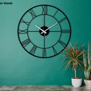 May include: A large, black metal wall clock with Roman numerals and silver hands. The clock is circular and mounted on a teal wall. The text "Silver Hands" is in the upper left corner. Decorative plants in white pots are in the foreground.