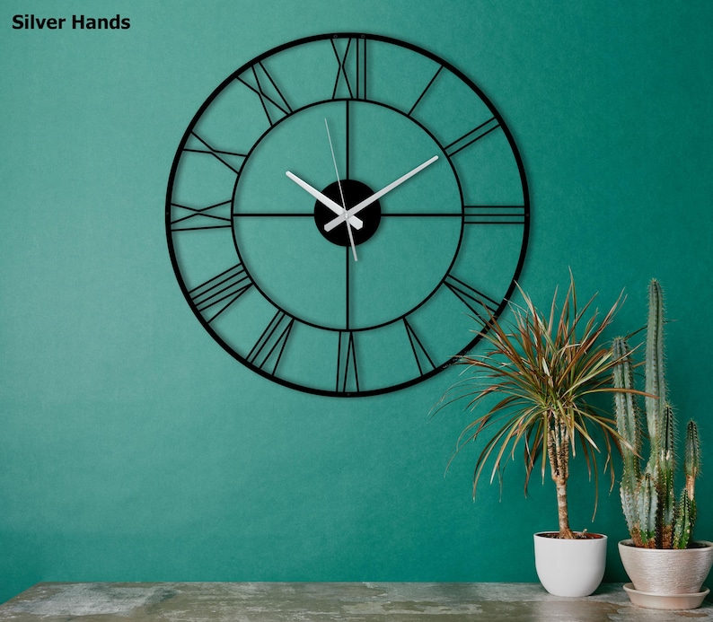 May include: A large, black metal wall clock with Roman numerals and silver hands. The clock is circular and mounted on a teal wall. The text "Silver Hands" is in the upper left corner. Decorative plants in white pots are in the foreground.