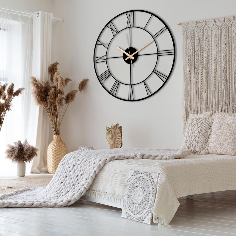 May include: A large, round black metal wall clock with Roman numerals. The clock hands are a light brown color. The clock is mounted on a white wall in a bedroom setting. The room has a neutral color palette with beige and white decor.