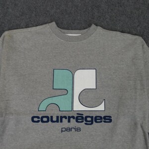 May include: A gray sweatshirt featuring a teal and white logo with the text "COURR&Egrave;GES Paris" in navy blue. The sweatshirt has a crew neck and a simple, classic design, suitable for casual wear.