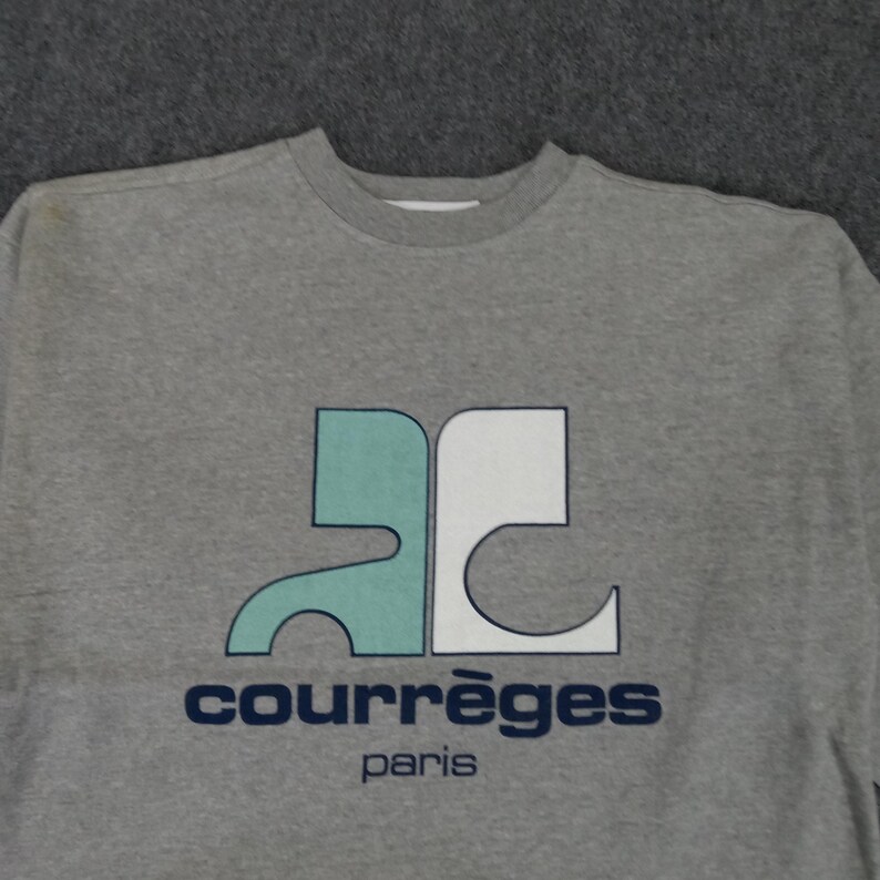 May include: A gray sweatshirt featuring a teal and white logo with the text "COURR&Egrave;GES Paris" in navy blue. The sweatshirt has a crew neck and a simple, classic design, suitable for casual wear.