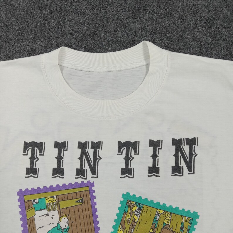 May include: White t-shirt featuring the word "TINTIN" in a bold, black font. Below the text are two colorful stamp-like illustrations. The shirt has a crew neck and short sleeves.
