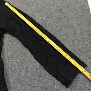 May include: A black long-sleeved garment sleeve positioned next to a yellow measuring tape. The tape shows the sleeve length is approximately 23.5 inches from the shoulder seam to the cuff edge, providing a clear visual reference for the size.